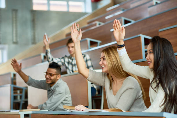 Students Raising Hands in Class Multi-ethnic group of students sitting at separate tables in lecture hall of modern college and raising hands, focus on two beautiful girls  smiling teenage high school girl raising hand during class stock pictures, royalty-free photos & images