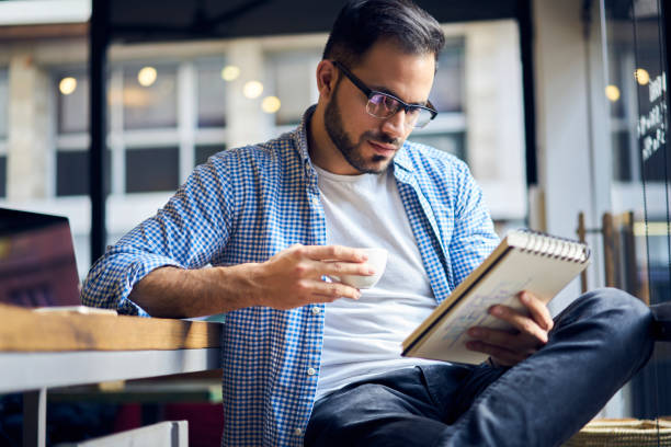istock Skilled handsome male chief editor reading script review of young author correcting mistakes in notebook while drinking coffee for breakfast during break preparing publication to print in newspaper 905464260