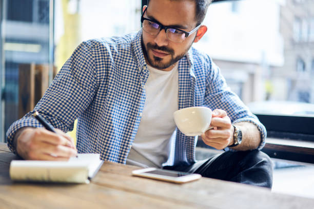 istock Talented handsome male blogger writing new article about traveling correcting mistakes and noting ideas into diary before publishing on webpage and social networks while working in modern cafe 905464270