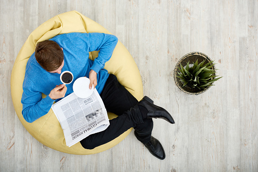 Directly above view of young man wearing blue sweater sitting on cozy beanbag chair, reading newspaper and enjoying fragrant coffee