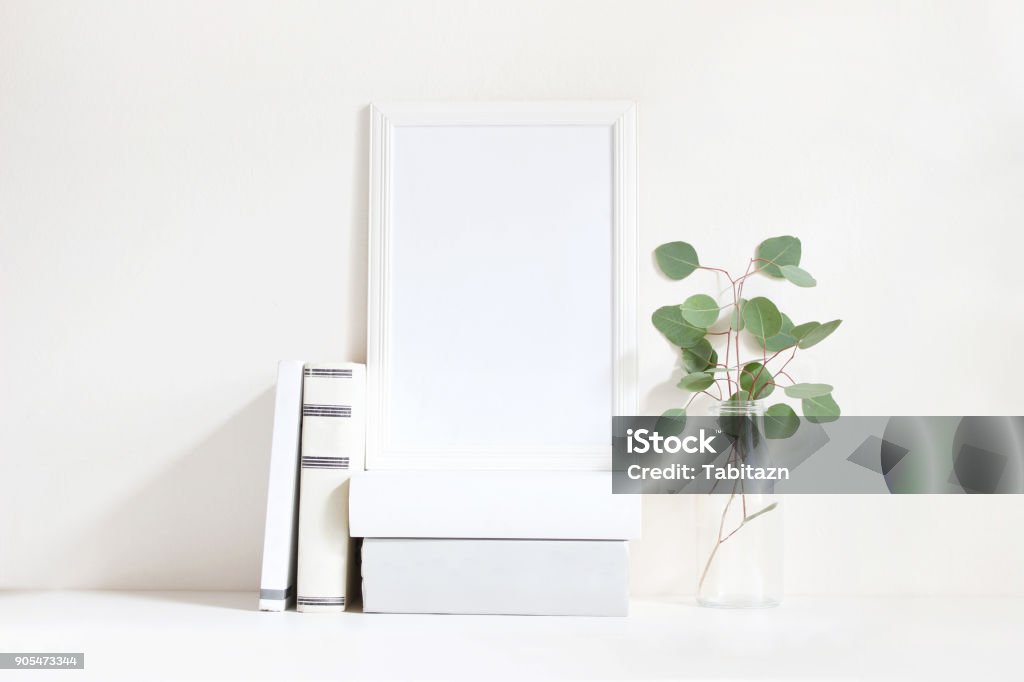 White blank wooden frame mockup with a green eucalyptus branches in glass bottle and pile of books lying on the table. Poster product design. Styled stock feminine photography. Home decor White blank wooden frame mockup with a green eucalyptus branches in glass bottle and pile of books lying on the table. Poster product design, styled stock feminine photography. Home decor. Border - Frame Stock Photo