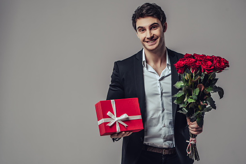 Handsome yound man in suit is posing on grey background with gift box and red roses in hands, looking at camera and smiling.