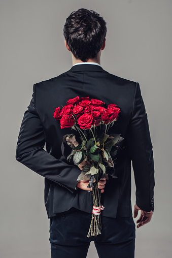 Handsome yound man in suit is standing with red roses behind the back on grey background.