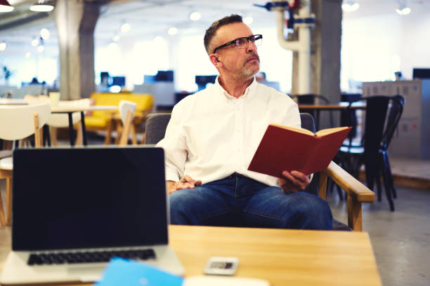 istock Mindful male executive thinking over plot of detective story reading favorite book while resting during break while waiting for email message using laptop computer and wireless connection in office 905481020