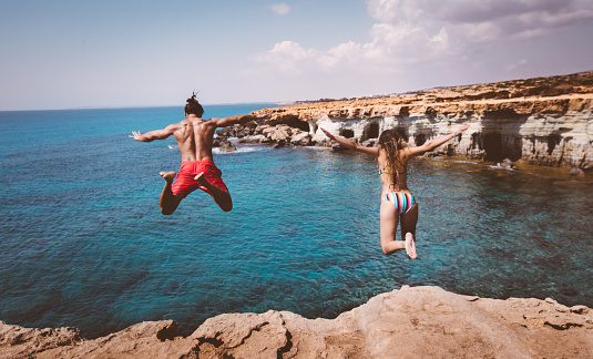 Young brave boyfriend and girlfriend jumping off cliff and diving into blue sea water