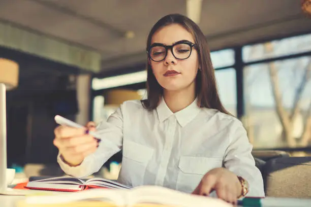 Photo of Portrait of concentrated skilled female literary editor working with articles for magazine correcting mistakes and editing composition while sitting at desk in modern coworking space with stationery