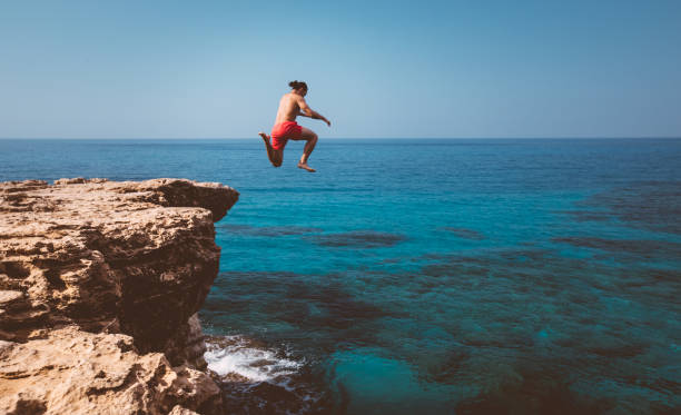 Young adventurous diver jumping off cliff into ocean Brave man diving from high cliff into blue sea waters on tropical island summer adventure people jumping sea beach stock pictures, royalty-free photos & images