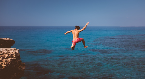 Young man jumping off cliff into tropical island blue waters in summer