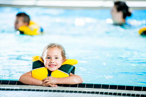 A Caucasian girl is taking a swimming class at a fitness center. She is in the swimming pool and wearing a life jacket. She is resting at the edge of the pool and smiling at the camera.