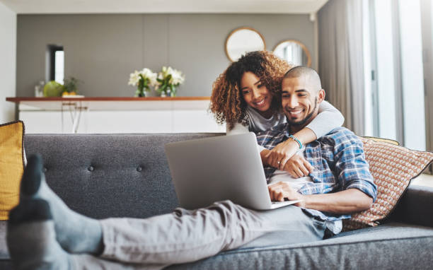 There's no denying their connection Shot of a young woman hugging her husband while he uses a laptop on the sofa at home young couple stock pictures, royalty-free photos & images