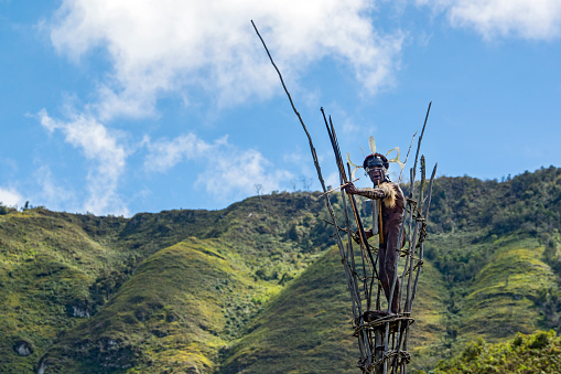 Baliem-Valley, New Guinea, Indonesia - August 22, 2017: The war chief of a small village of the Dani  people (also spelled as Ndani) on the observation tower of the village. He is traditionally dressed and wearing also the typical Koteka (penis sheath or gourd) and is holding a bow in his hand. The Dani inhabit the Baliem Valley in the central highlands of West Papua (the Indonesian part of New Guinea).