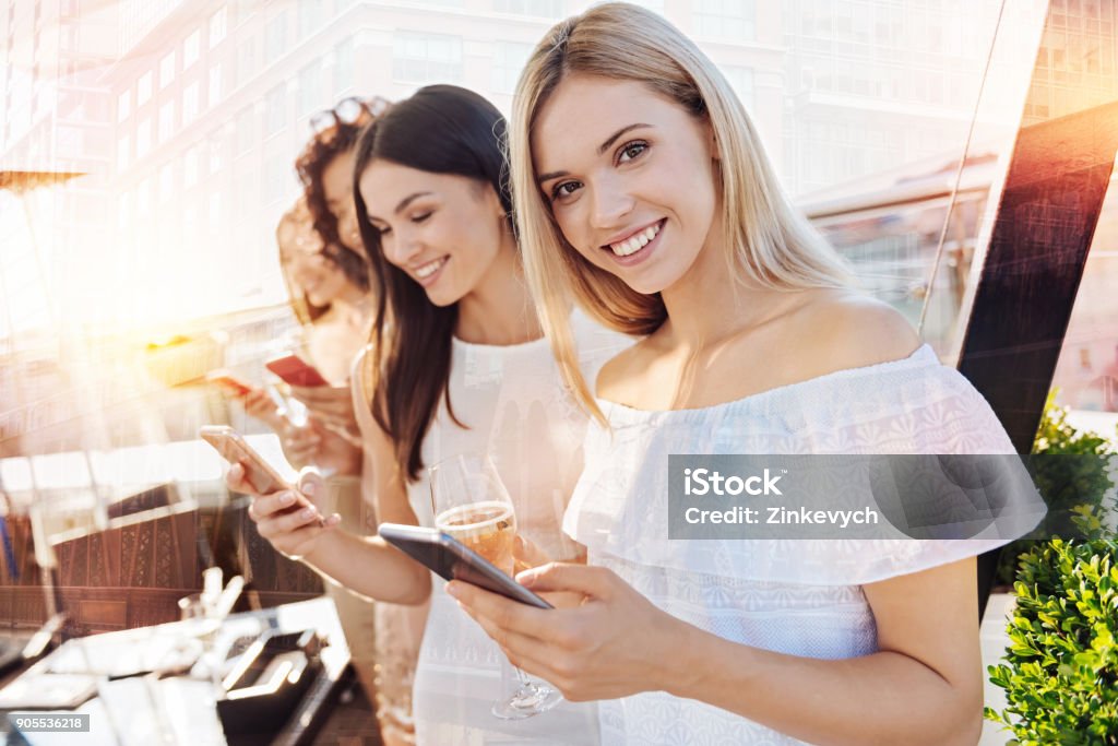 Joyful females holding their phones Look at me. Attractive blonde keeping smile on her face and bowing head while looking straight at camera Bachelorette Party Stock Photo