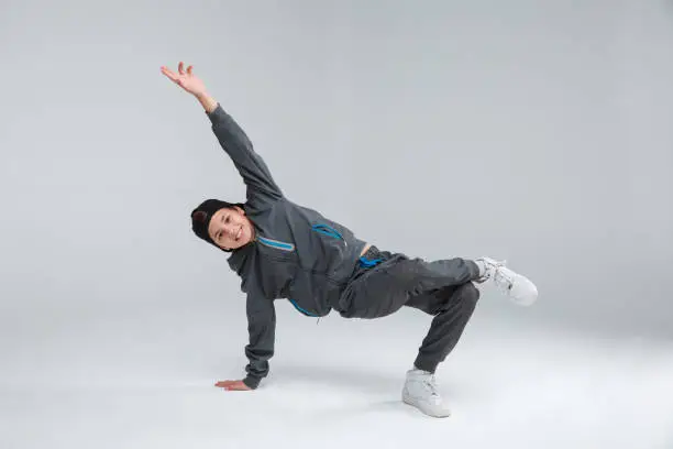 Photo of A cheerful little boy dressed in a gray tracksuit, actively dancing standing on one arm and one leg. Gray background.