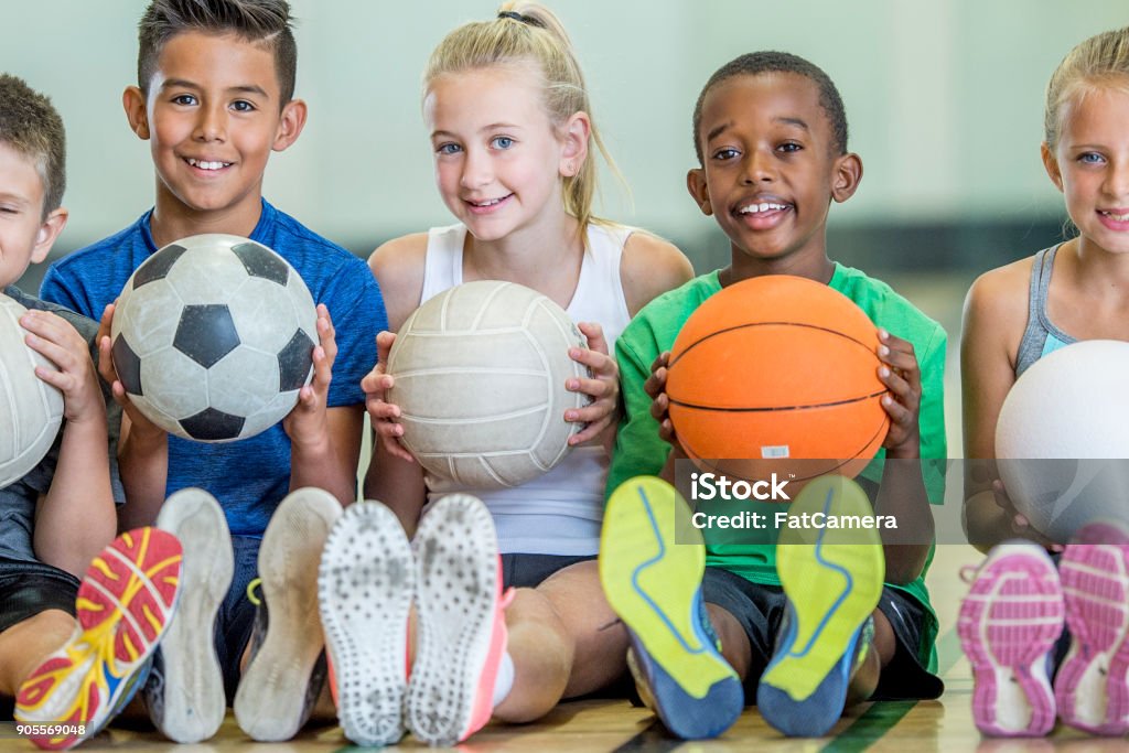 Physical Education A group of children are indoors in their elementary school gym. They are sitting on the floor and getting ready to play sports. The kids are holding a basketball, a soccer ball, and three volleyballs. Child Stock Photo