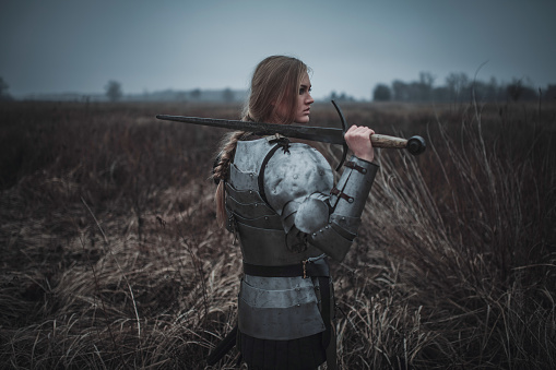 Girl in image of Jeanne d'Arc in armor and with sword in her hands stands on meadow in middle of dry grass. Back view.