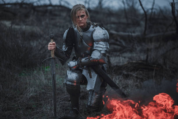 Girl in image of Jeanne d'Arc in armor and with sword in her hands kneels against background of fire and smoke. Girl in image of Jeanne d'Arc in armor and with sword in her hands kneels on meadow against background of fire, black smoke and dry grass. sword photos stock pictures, royalty-free photos & images