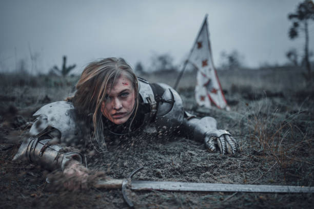 dziewczyna na zdjęciu jeanne d'arc w zbroi czołga się w błocie z mieczem w rękach na tle flagi. - jeanne zdjęcia i obrazy z banku zdjęć