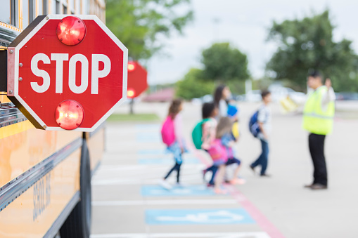 Focus is on a flashing school bus stop sign in the foreground as a group of schoolchildren cross a parking lot with the help of a crossing guard in the distance.