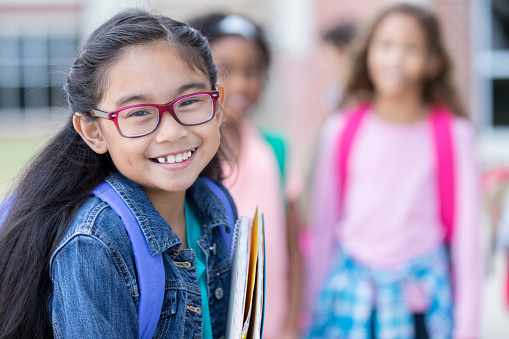 Focus is on an adorable little elementary age girl as she smiles for the camera outside her school on the first day.  Other classmates can be seen in the background.