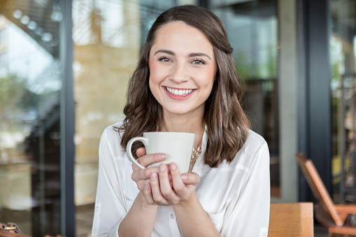 Attractive young woman enjoys relaxing at an outdoor cafe. She is smiling while holding a hot beverage.