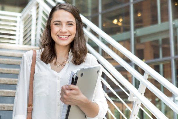 Confident college student on her way to class Attractive Hispanic female college student holds books and a laptop while walking to class on the university campus. A staircase is in the background. adult education book stock pictures, royalty-free photos & images
