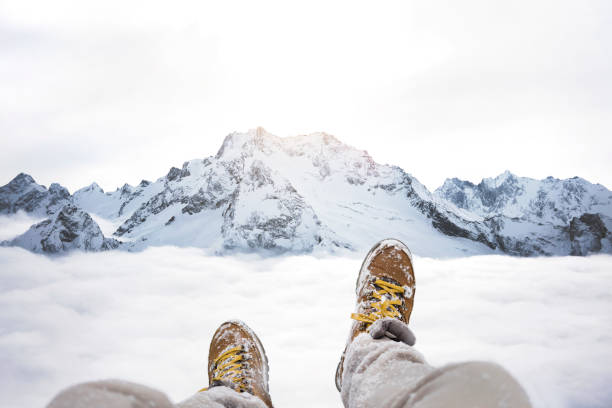 voyageur assis sur le sommet de la montagne, vue pov sur grand hiver montagne au-dessus du nuage et de bottes de randonnée. jambes de close up sur fond de paysage de roche neigeux - climbing rock climbing rock mountain climbing photos et images de collection