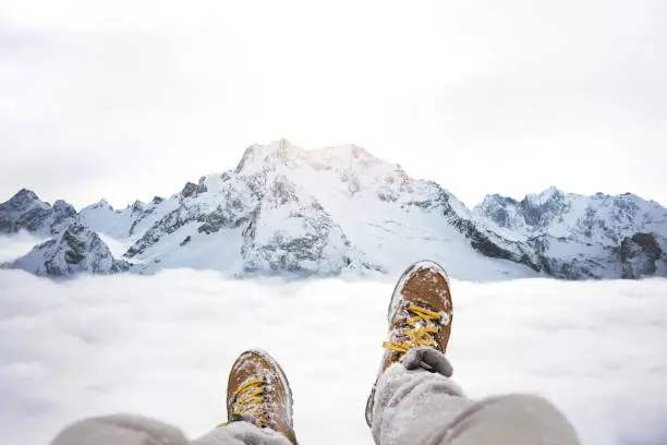 Traveler sitting on mountain peak, POV view on great winter mountains above the cloud and hiking boots. Legs of close up on background of rock snowy landscape