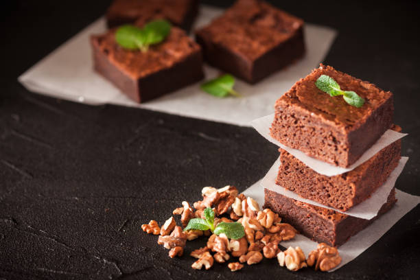 istock Chocolate brownie square pieces in stack on white plate with walnuts, decorated with mint leaves and cocoa on black background. Delicious dessert. Dark mood. Close up photography. Selective focus 905643416