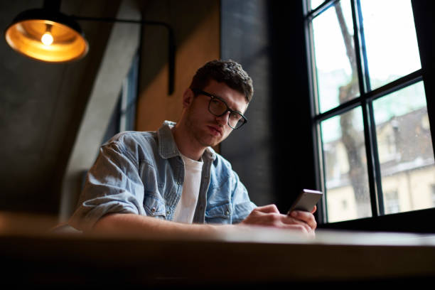 istock Portrait of confident hipster guy in optical eyeglasses looking at camera while chatting online in social networks on mobile phone sitting in university interior.Copy space area for advertising 905653830
