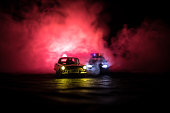 Toy BMW Police car chasing a Ford Thunderbird car at night with fog background. Toy decoration scene on table . Selective focus 