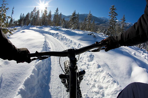 The handlebar view of a man riding a winter fat bike on a groomed trail. The bike has a front suspension fork and extra wide tires for riding on the snow.