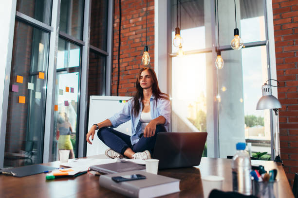 young businesswoman meditating sitting on working table in lotus pose in her office - yoga business women indoors imagens e fotografias de stock