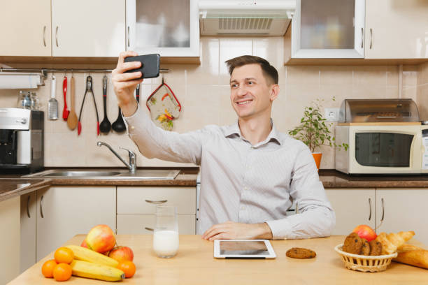 istock Handsome young business man in plaid shirt having breakfast, sitting at table, doing selfie on mobile phone, eating cereals with milk on light kitchen. Healthy lifestyle. Cooking at home. Prepare food 905742496