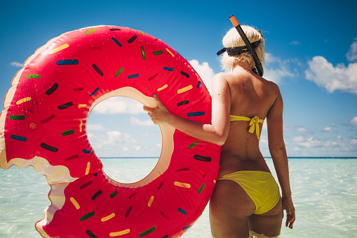 Attractive and very slim woman is standing in shallow water. She have whole equipment for snorkeling, tube and glasses. With her left hand she is holding on inflatable float. This float have shape of doughnut witch is missing one part, something like one bite. Her short blonde hair is wet so she is just from water. Water is crystal clear and cloudy sky is picturesque but there is no worry about rain. Peaceful environment and heavenly island is amazing combination for enjoyment and relaxing.