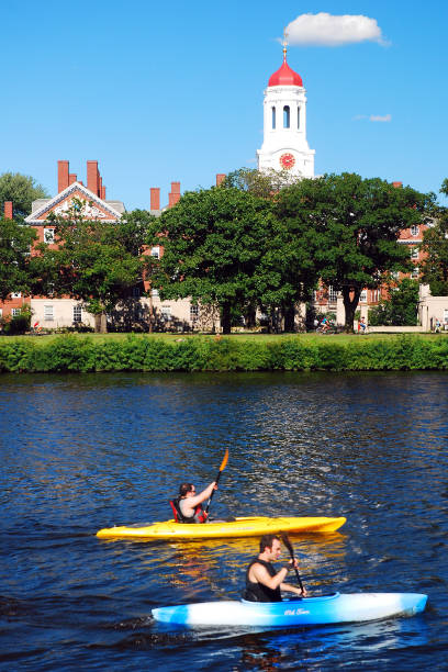 Kayakers Pass Harvard University on the Charles River Cambridge, MA, USA August 11, 2011 Two Kayakers on the Charles River in Cambridge, Massachusetts pass the red topped Dunster House, a recognizable landmark on the Harvard University campus charles river stock pictures, royalty-free photos & images