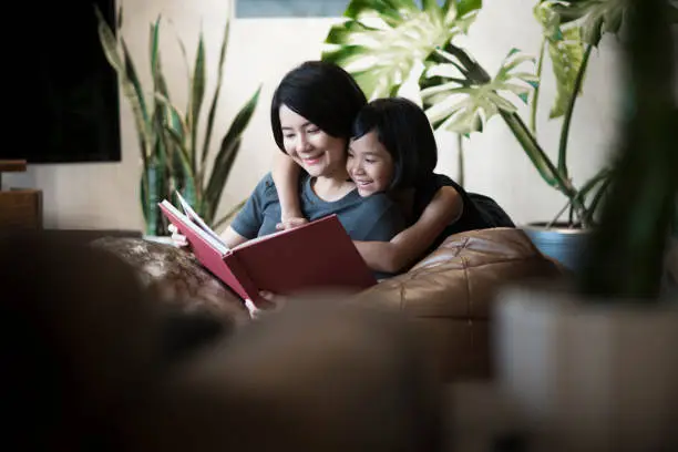 Photo of Asian mother and daughter reading a book at home.