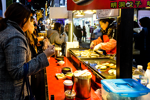 The Gwangjang Market in Seoul is full of street food vendors cooking and preparing food, while hungry patrons happily eat and enjoy. Photo taken during a cold winter evening.