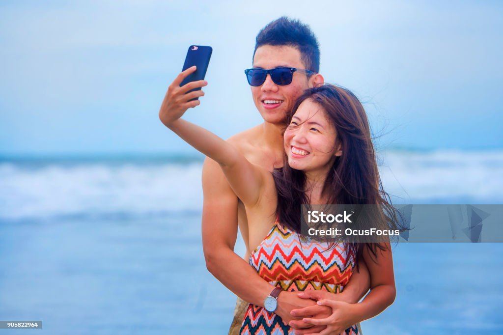 young happy and beautiful Asian Chinese couple taking selfie photo with mobile phone camera smiling joyful having fun on the beach in romance and lovers holidays trip picture concept Beach Stock Photo