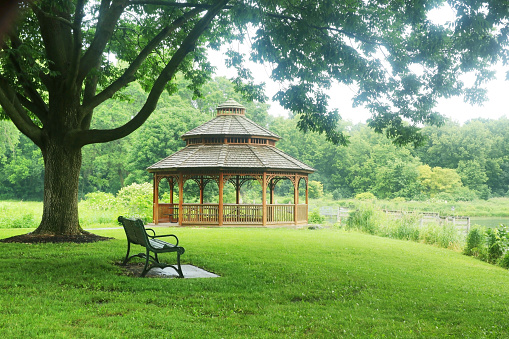 Summer landscape in a city park with a bench on a foreground and wooden gazebo on a background during warm rainy day.