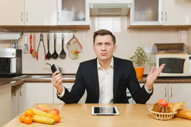 Photo of Perturbed young business man in suit, shirt, having breakfast, sitting at table with tablet, spreading hands with mobile phone, eating fruits on light kitchen. Healthy lifestyle. Cooking food at home.