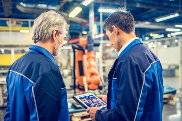 Two engineers examining robotic arm in factory Rear view of two engineers looking at a digital tablet while standing in front of a robotic arm. computer aided manufacturing stock pictures, royalty-free photos & images