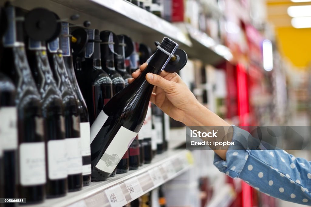 La donna sta comprando una bottiglia di vino sullo sfondo del supermercato - Foto stock royalty-free di Alchol