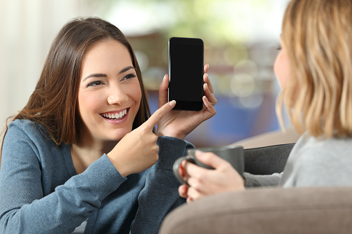 Happy girl showing a blank phone screen to a friend on a couch at home