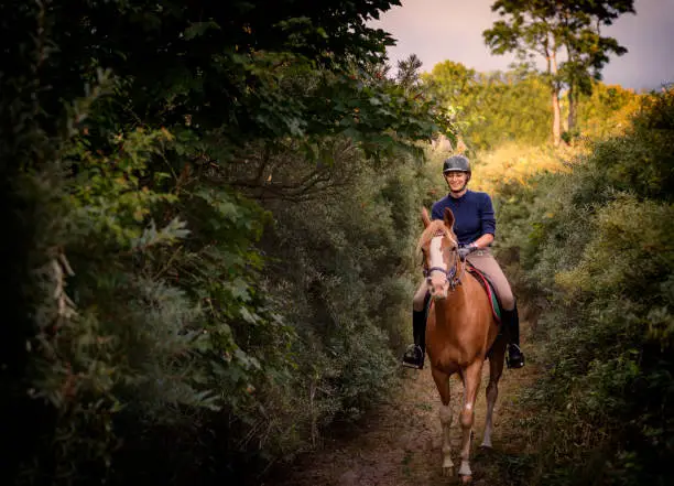 Portrait of a young woman who is riding her tan coloured Arabian horse through the woods on her way to the beach. Photographed the island of Møn in Denmark. Colour, horizontal with some copy space.