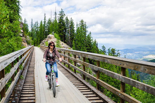 Young Woman Riding a Bicycle on a Wooden Testle Bridge on a Cloudy Spring Day. Myra Canyon, Kelowna, BC, Canada.