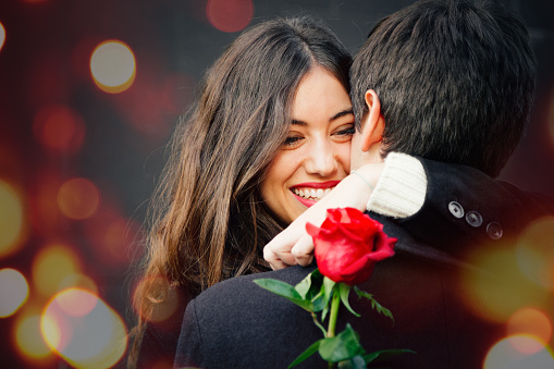 Pretty and happy couple in love cuddling on the street holding a red rose. Lights bokeh effect.