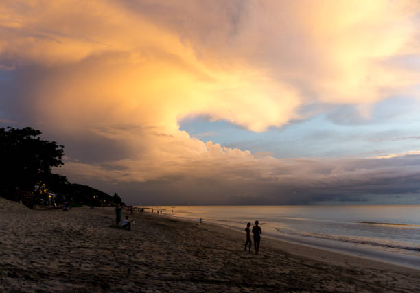 Numerous stratus, cumulus, and stratocumulus clouds are layered across the sky.  These are low level clouds typically associated with fair weather.  The location is a beach on Ko Lanta, Krabi, Thailand, during Summer which is Monsoon Season.  Several people are admiring the beautiful sunset scene.