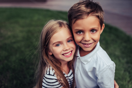 Couple of charming childs having fun outdoors. Portrait of beautiful girl and boy are hugging, smiling and looking at the camera. Little brother and sister in park on a green grass.