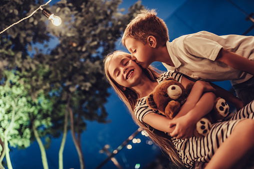 Kids are playing in the evening in park with garland of light bulbs. Charming boy and girl are enjoying spending time together. Brother and sister are sitting on a grass with toy bear in hands. Children's first kiss