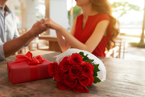 Unrecognizable young couple holding hands while having romantic date at lovely outdoor cafe, bouquet of red roses and gift box lying on wooden table